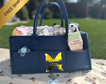 May include: A navy blue diaper bag with a gold butterfly and the name "Olivia" on the front. The bag has multiple pockets and is filled with baby supplies.