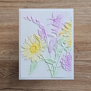 May include: A white greeting card with a floral design. The embossed flowers include a large yellow daisy, lavender gladiolus, and a small purple flower. The card is set against a wooden background.