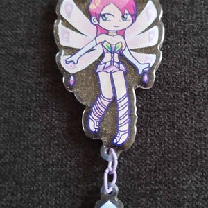 Peut inclure: Un porte-clés en acrylique transparent représentant une fée de dessin animé aux cheveux roses, aux ailes et à une robe. Le porte-clés a un fond doré pailleté et un charme d'une bague avec une gemme bleue.