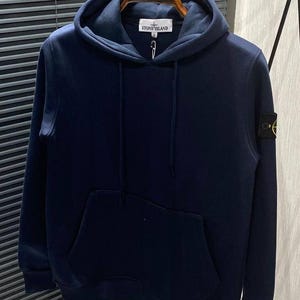 Könnte beinhalten: Marineblauer Kapuzenpullover mit Kängurutasche und Kapuze mit Kordelzug. Der Pullover hat einen kleinen Aufnäher am linken Ärmel und ein Logo auf der Brust. Er hängt an einem Holzbügel.