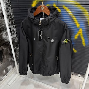 Puede incluir: Una chaqueta negra con capucha que presenta un cierre de cremallera frontal, dos bolsillos laterales con cremallera y una capucha con cordón. Una etiqueta de Stone Island cuelga del cuello y un parche de brújula amarillo y verde en la manga.