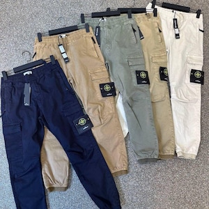 Può includere: Un'esposizione di pantaloni cargo in vari colori: blu navy, marrone, verde oliva, kaki e bianco sporco. Ogni paio presenta una tasca cargo e una toppa del marchio. I pantaloni sono appesi a grucce nere.