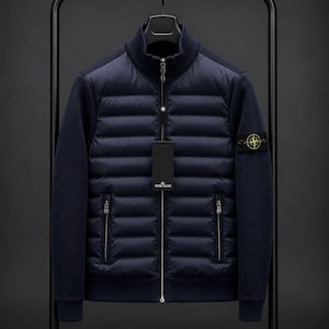 Könnte beinhalten: Eine marineblaue Hybridjacke mit gestepptem, wattiertem Vorderteil, durchgehendem silbernem Reißverschluss und zwei seitlichen Reißverschlusstaschen. Die Ärmel und der Rücken bestehen aus geripptem Strickstoff. Ein Stone Island Logo-Patch ist am linken Ärmel.