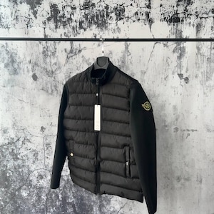 Puede incluir: Una chaqueta acolchada negra con cuello alto, cremallera frontal completa y mangas largas. La chaqueta presenta un parche circular bordado en la manga izquierda y una etiqueta rectangular blanca que cuelga del tirador de la cremallera.