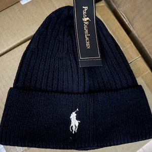 Può includere: Un berretto in maglia a coste blu navy con bordo ripiegato. Il berretto presenta un logo di giocatore di polo bianco ricamato sul davanti e un'etichetta nera con "Polo Ralph Lauren" stampato in oro.