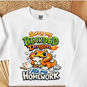 以下が含まれることがあります： 宿題を食べている漫画のヒゲトカゲのカラフルなグラフィックが特徴の白いスウェットシャツ。「Sorry my Bearded Dragon Ate my Homework」の文字が緑、オレンジ、青で書かれています。