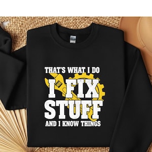 May include: Black sweatshirt with the text "THAT'S WHAT I DO I FIX STUFF AND I KNOW THINGS" in white and yellow. The design includes a wrench and gear graphic. The sweatshirt is displayed on a woven surface.