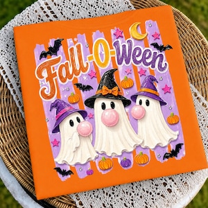 Könnte beinhalten: Ein orangefarbenes T-Shirt mit dem Schriftzug Fall-O-Ween in orangefarbener und violetter Schreibschrift. Das Motiv zeigt drei weiße Geister mit violetten und schwarzen Hexenhüten, die jeweils eine rosa Kaugummiblase blasen, umgeben von Fledermäusen, Kürbissen und Sternen.