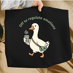 May include: Black tote bag featuring a cartoon goose with a coffee cup and a green messenger bag. The goose is walking and the text "Off to regulate emotions" is arched above the goose in green.