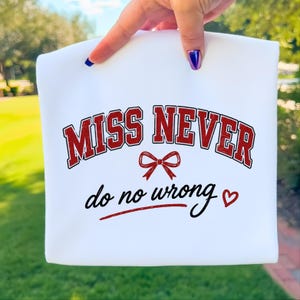 May include: A white t-shirt featuring the red text MISS NEVER in a collegiate font, a red bow graphic, and the black cursive text do no wrong with a red heart symbol. The design is centred on the front of the garment against a plain white background.