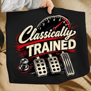 Może przedstawiać: Czarny t-shirt z nadrukiem graficznym przedstawiającym obrotomierz samochodowy z czerwoną wskazówką, trzy metalowe pedały oraz napis Classically TRAINED kremowymi i czerwonymi literami. Projekt podkreśla elementy sterowania manualną skrzynią biegów.