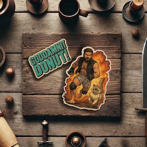 May include: Two stickers, one with a cartoon of a bearded man running with a cat, and the other with the text "GODDAMNIT DONUT!" in a retro style.