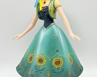 May include: A figurine of a character from the movie Frozen, wearing a teal dress with sunflower details. The figure has red hair, a yellow top, and a green vest. The figurine is standing on a green base.