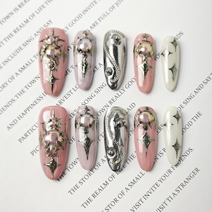 Puede incluir: Una selección de diez uñas artificiales con diversos diseños. Las uñas presentan colores como rosa, plata y blanco, y están adornadas con adornos metálicos como estrellas, perlas y detalles de cadena. Las uñas están dispuestas sobre una superficie blanca con texto impreso.