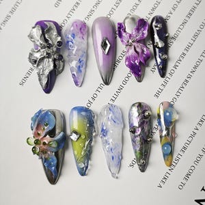 May include: A collection of ten long, almond-shaped artificial nails. Each nail features unique designs, including floral embellishments, gems, and abstract patterns in shades of purple, blue, green, and silver. The nails are arranged on a white surface.
