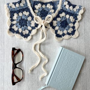 Può includere: Un colletto all'uncinetto fatto a mano nei toni del blu e del bianco, con un motivo a granny square e una chiusura a fiocco color crema. Sono presenti anche occhiali tartarugati e un diario azzurro con motivi floreali in rilievo.