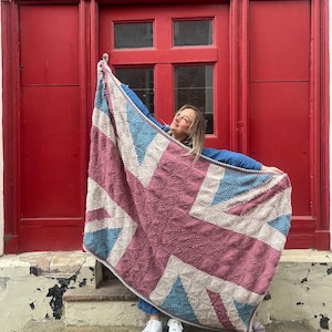 May include: A person holding a large crocheted blanket with a Union Jack design in pink, blue, and white. The blanket is draped over the person's shoulders and they are smiling.