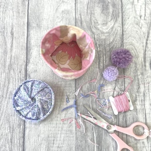 May include: A fabric storage basket with a pink floral interior, a matching circular fabric lid with a lavender floral pattern, two purple yarn pom-poms, a spool of pink embroidery thread, and a pair of pink-handled scissors with star accents.