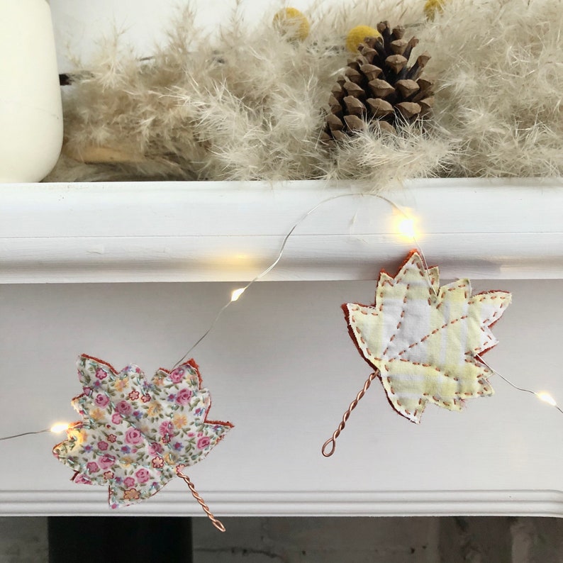 May include: A string of fairy lights with fabric leaf decorations. The leaves are made of different patterned fabrics, including floral and striped patterns. The lights are strung on a white surface.