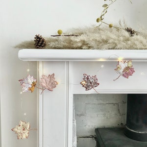 May include: A white fireplace mantel with a string of lights decorated with fabric fall leaves. The leaves are in shades of pink, white, and brown with floral patterns.