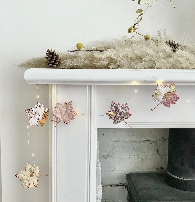 May include: A white fireplace mantel with a string of lights decorated with fabric fall leaves. The leaves are in shades of pink, white, and brown with floral patterns.
