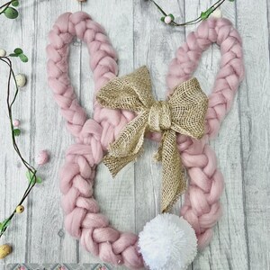 May include: A pink braided wool bunny wreath with a hessian bow and a white pom-pom tail. The wreath is decorated with faux greenery and Easter eggs. The text "get Smitten" is visible at the bottom.