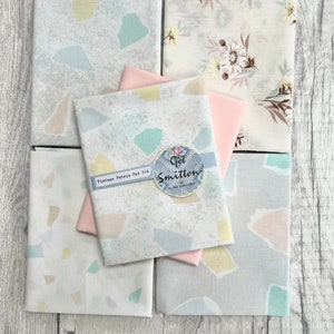 May include: A set of five fabric fat quarters in a pastel colour palette. The fabrics feature a variety of patterns, including a floral print, a geometric print, and a solid pink fabric. The fabrics are labelled "Vintage Fabric Fat 1/4" and "Get Smitten by Lisa Congdon".