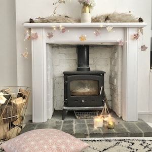 May include: A white fireplace mantel with a black wood-burning stove. The mantel is decorated with a garland of fall leaves and a vase of pink flowers. There are two candles on the hearth in front of the stove.