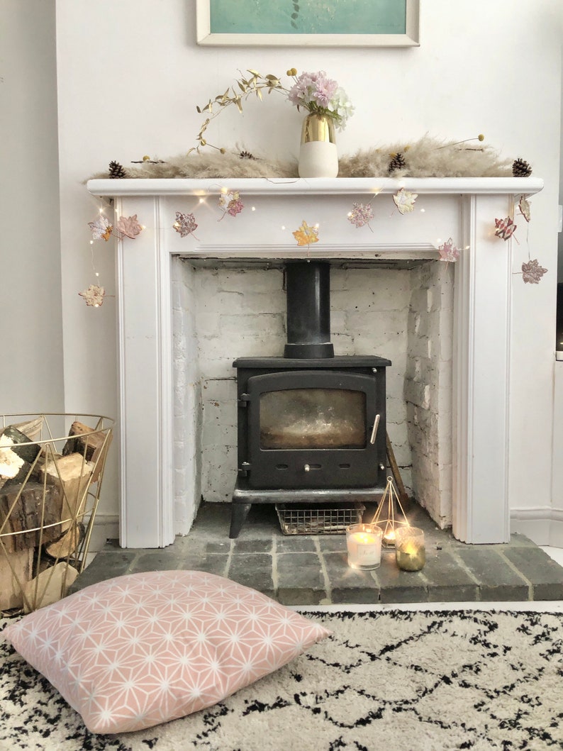 May include: A white fireplace mantel with a black wood-burning stove. The mantel is decorated with a garland of fall leaves and a vase of pink flowers. There are two candles on the hearth in front of the stove.