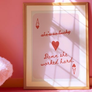 May include: A framed print of a pink playing card with a red ace of hearts and the text "she's so lucky" crossed out and "Damn she worked hard." printed below.