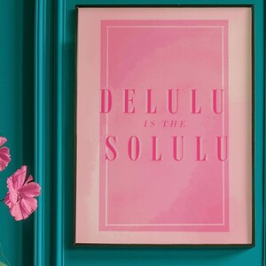 May include: A framed pink art print with the words "DELULU IS THE SOLULU" in a bold, serif font. The print is set against a light pink background and framed in a black frame. The wall behind the frame is teal.