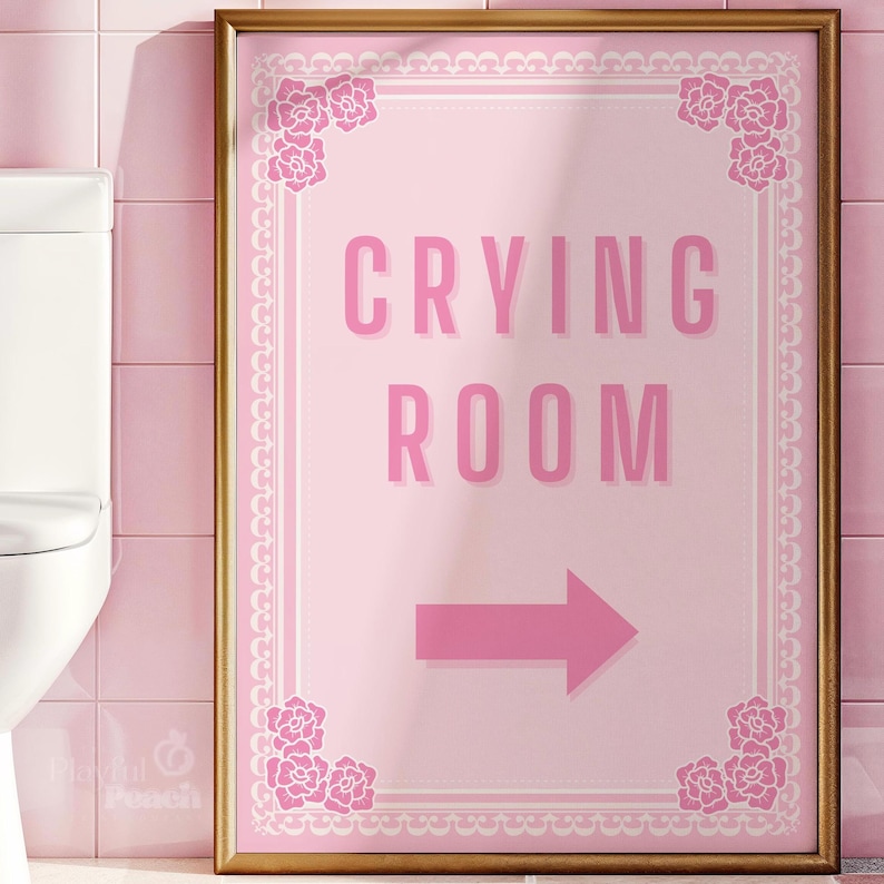 May include: A pink sign with a gold frame and white floral accents. The sign reads "Crying Room" with an arrow pointing to the right.