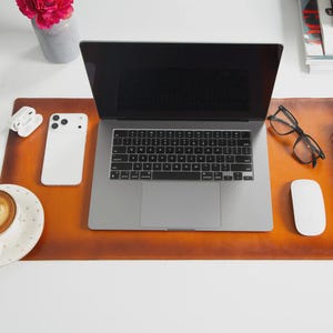 Könnte beinhalten: Ein Arbeitsplatz mit einem Laptop, Smartphone und kabellosen Ohrhörern auf einer braunen Leder-Schreibtischunterlage. Eine Tasse Kaffee mit Latte Art, eine Brille und ein Notizbuch sind ebenfalls vorhanden. Eine Vase mit rosa Blumen befindet sich in der Nähe.