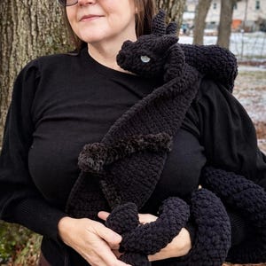 May include: A black crocheted dragon plushie with a fluffy mane and a light blue eye. The dragon is wrapped around the person's shoulders and arms. The plushie is made of a soft, textured yarn.