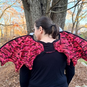 May include: A pair of handmade crochet wings in shades of pink and black, with a black border. The wings are designed to be worn, with a neckline that fits around the shoulders. The wings are displayed outdoors.