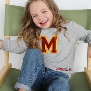 May include: A young person wearing a gray sweatshirt with a large maroon and gold letter "M" and the text "MINNESOTA KID". They are sitting in a wooden director's chair with a green canvas seat and back, smiling at the camera.