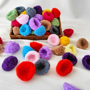 Op de afbeelding: Een verzameling kleurrijke gehaakte haar elastiekjes in verschillende kleuren, waaronder roze, paars, blauw, groen, rood, geel en oranje. De elastiekjes zijn gerangschikt in een mand en op een wit oppervlak.