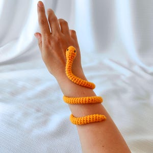 Könnte beinhalten: Ein gehäkeltes orangefarbenes Schlangenarmband, das sich um einen Arm wickelt. Die Schlange hat einen detaillierten Kopf mit schwarzen Augen und windet sich um das Handgelenk und den Unterarm. Das Armband besteht aus strukturiertem Garn.
