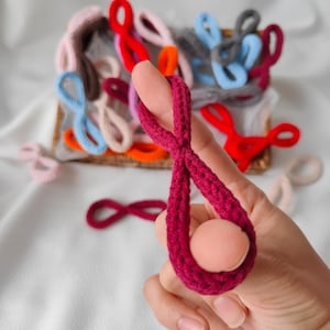Op de afbeelding: Een hand houdt een bordeauxrood gehaakt item vast met een perzikkleurige bol. Andere gehaakte items in verschillende kleuren, waaronder rood, blauw en roze, zijn op de achtergrond te zien. De items lijken handgemaakt.
