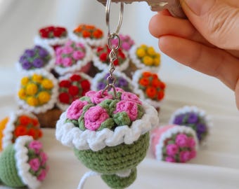 Puede incluir: Un llavero de flores de ganchillo con flores rosas en una maceta verde con un borde blanco de ganchillo. El llavero está unido a un llavero plateado.