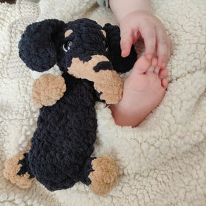 Könnte beinhalten: Ein handgefertigtes Dackel-Spielzeug aus Plüsch in Schwarz und Hellbraun. Das Spielzeug besteht aus weichem, strukturiertem Garn und hat Schlappohren und kleine, runde Augen. Der Hund liegt auf einer cremefarbenen, flauschigen Decke.