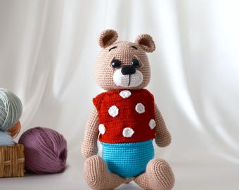 May include: A crocheted teddy bear wearing a red shirt with white polka dots and blue pants. The bear is sitting on a white surface.