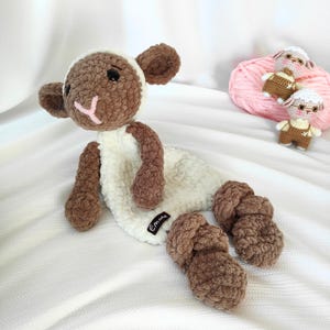May include: A handmade crochet lamb doll with a white body and brown head, ears, arms, and legs. The lamb has a pink mouth and black eyes. A small tag with the name "Emma" is attached. Two smaller lamb dolls and a ball of pink yarn are in the background.