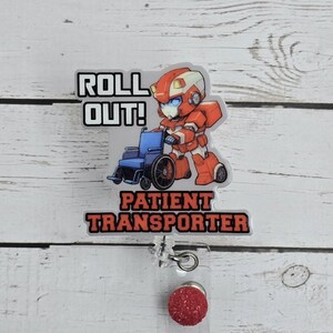 May include: A badge reel with a red and orange robot pushing a blue wheelchair. The text "ROLL OUT!" is above the robot, and "PATIENT TRANSPORTER" is below. The reel has a clear plastic base and a red glitter button.
