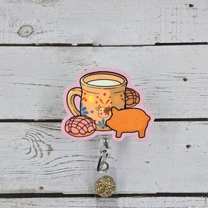 May include: A badge reel featuring a decorative illustration of a tan mug filled with white liquid, adorned with a small cow face and colorful floral patterns. Beside the mug are two round, textured pastries and an orange pig-shaped cookie.