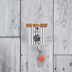 May include: A white badge reel with a cartoon duck wearing a black and white striped prison uniform behind bars. The badge reel has the text "GOT TOO SILLY" in orange letters. An orange pom-pom is attached to the reel.