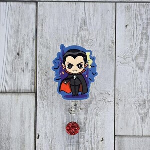 May include: A badge reel featuring a cartoon vampire character with a black suit, red cape, and a blue background with bats and a castle. The reel has a clear plastic body and a red glitter accent.