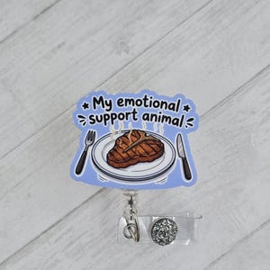May include: A badge reel featuring a light blue background with a graphic of a grilled steak on a white plate, a fork, and a knife. The text reads My emotional support animal. The reel has a clear plastic strap and a circular silver glitter accent.