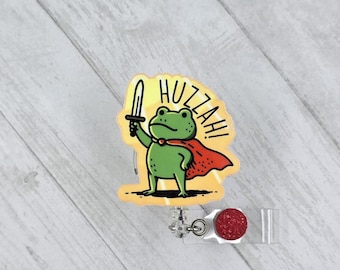 May include: A badge reel featuring a green frog wearing a red cape and holding a sword, with the text HUZZAH! printed above the frog. The badge reel has a circular red glitter accent and a clear plastic strap attachment.