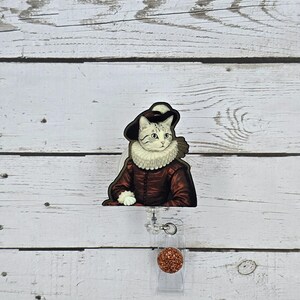 May include: A badge reel featuring a cat dressed in Renaissance attire, including a dark hat and a ruffled collar. The cat is depicted in a maroon coat. The reel has a clear plastic base and a round, glittery, copper-colored accent.